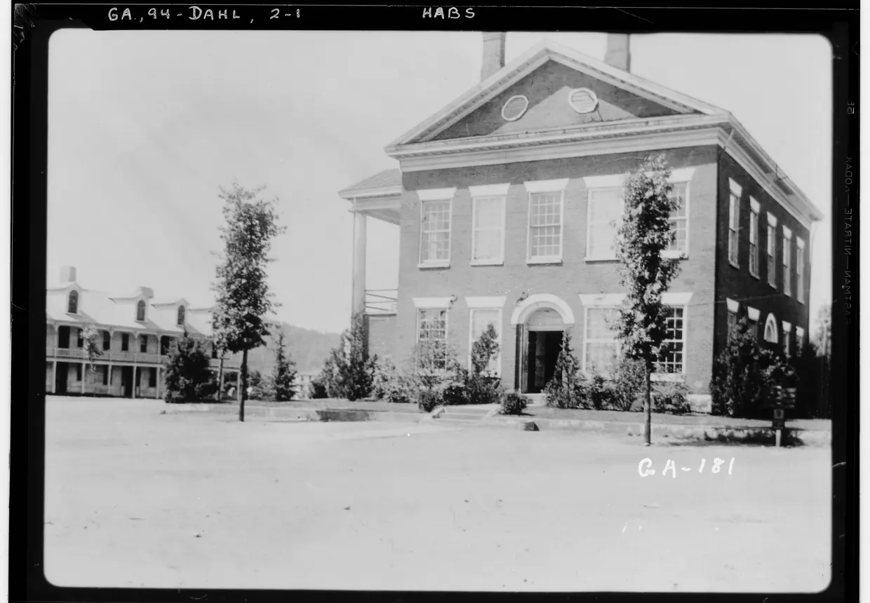 A black-and-white view of a two-storey brick courthouse with a pedimented gable, two round attic windows, chimneys and a side portico, young trees on a bare dirt square.
