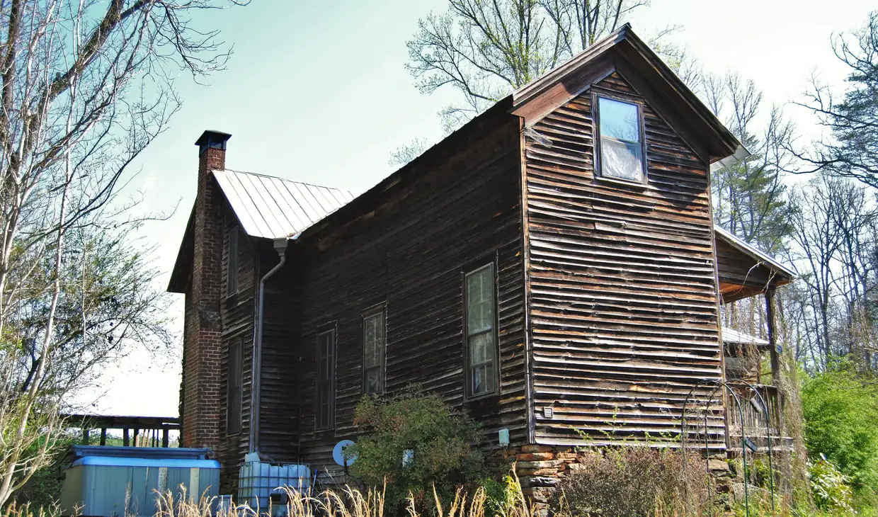 A tall weathered board-sided farmhouse with a metal roof, a chimney at the far end and a small porch, seen from a garden of tall grass and shrubs under bare spring trees.