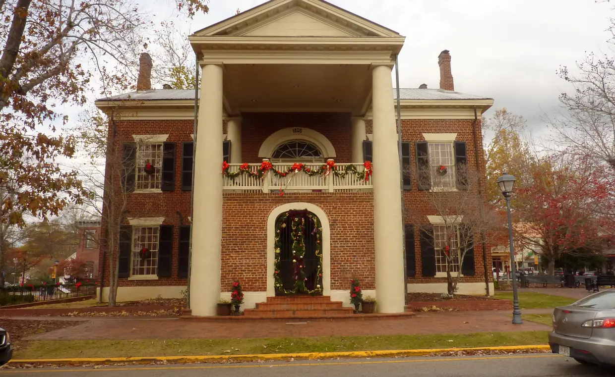 A columned brick building front with an arched doorway and a balcony rail, windows hung with green wreaths and red bows, autumn trees on both sides and a lamp post at the curb.