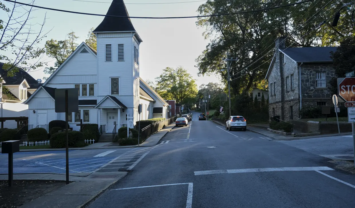 A quiet street crossing in late-day light: a white wooden church with a pointed steeple on the left, a grey stone building behind a stop sign on the right, big trees along the street.