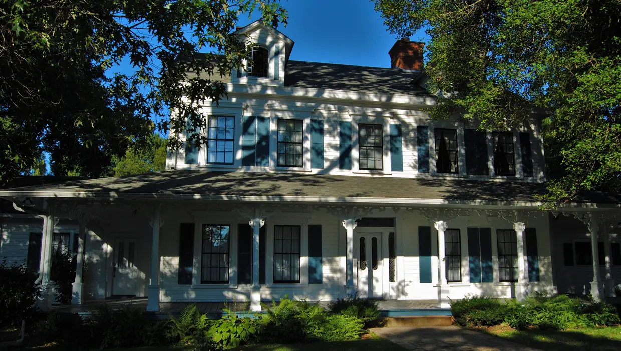 A large white two-storey house with a long columned wrap-around porch, black shutters, dormers and a brick chimney, half in tree shade behind a lawn and shrubs.