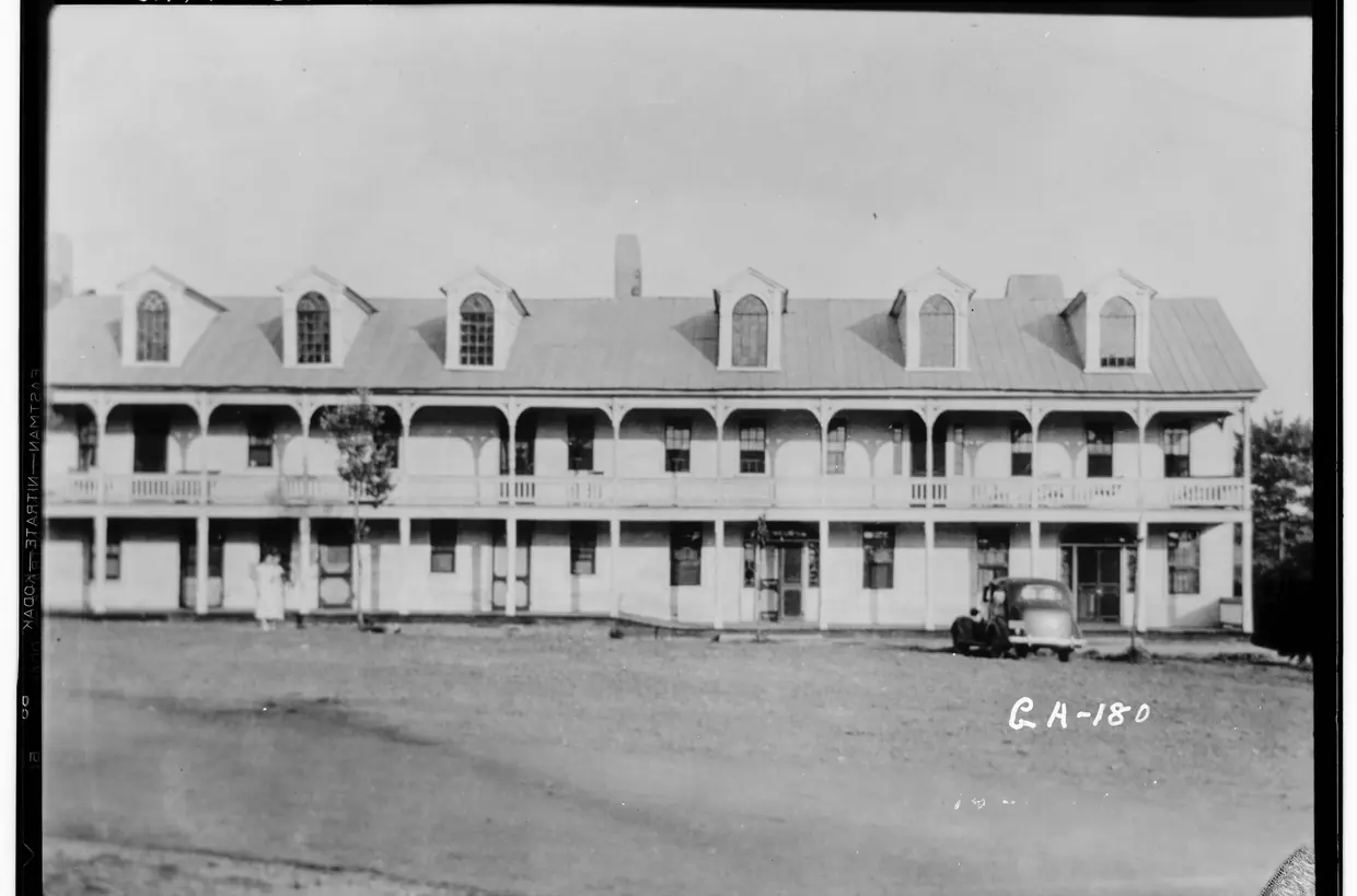 A black-and-white view of a long two-storey building with a row of dormers in its metal roof and porches on both floors, a 1930s car parked on a bare dirt square.