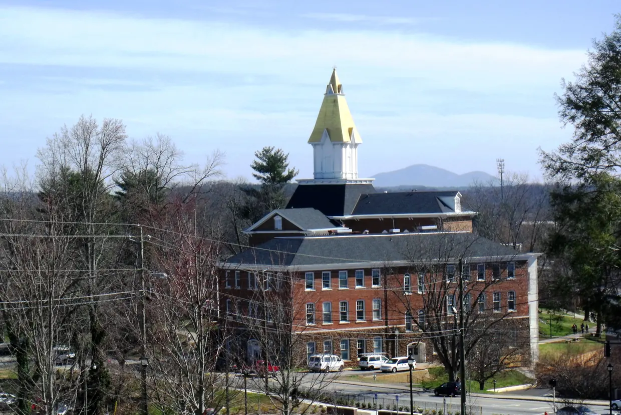 A gold-coloured steeple above a red-brick hall with a dark mansard roof, seen across a campus street and lawn, with a blue mountain on the horizon.