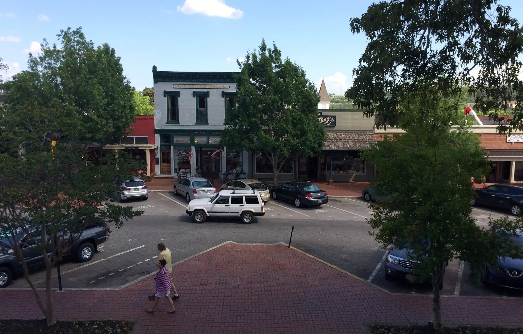 A row of two-storey shopfronts with awnings across an angle-parked lot, a brick plaza in front with two people walking past young trees, under a summer sky with small clouds.