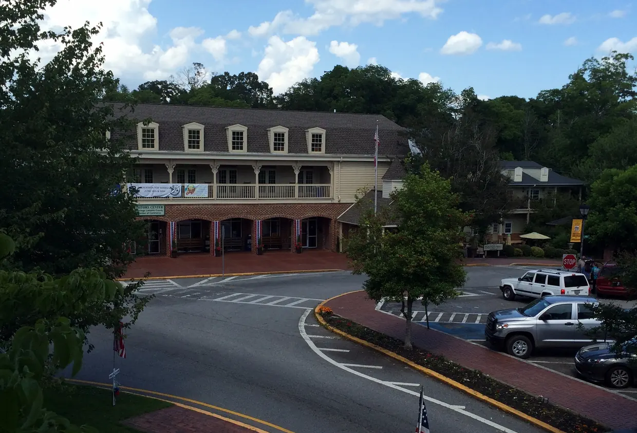 A long two-storey building with a row of dormer windows and a full-length upper porch facing a curving street, parked cars and a brick-edged lawn with an American flag, wooded hills behind.