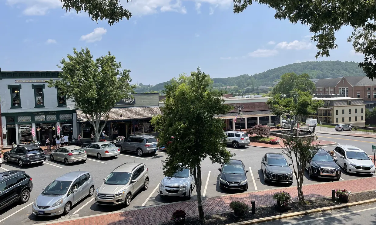 A high view over a parking lot of cars to a row of low shopfronts under shade trees, a brick multi-storey building at the right and a green wooded hill under a blue sky.