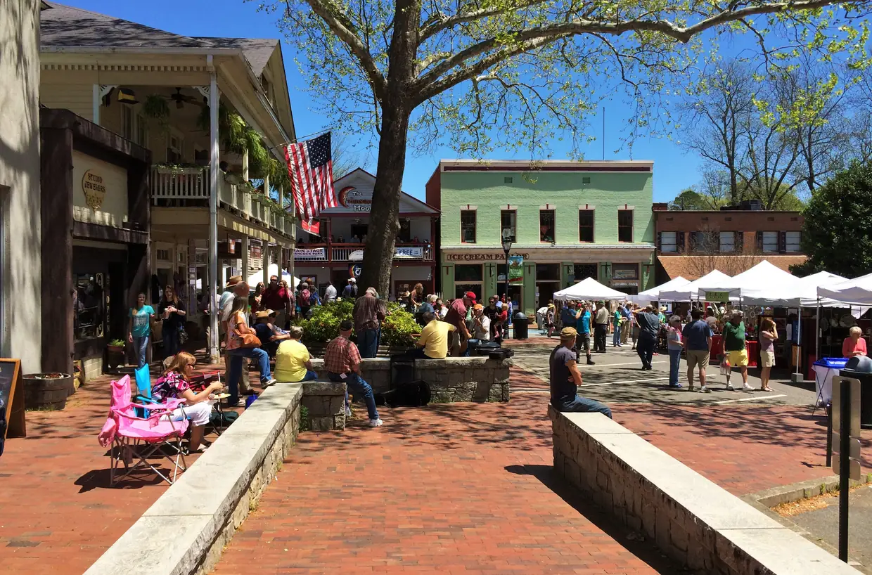 A busy brick sidewalk under a large shade tree, white vendor tents along the far side, a covered porch with an American flag and a pale-green two-storey storefront behind.