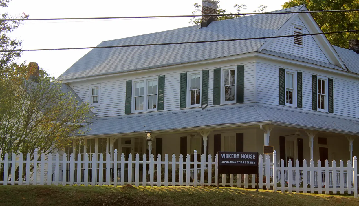 A two-storey white clapboard house with dark-green shutters, brick chimneys and a deep wraparound porch behind a white picket fence, with a sign reading Vickery House, Appalachian Studies Center.